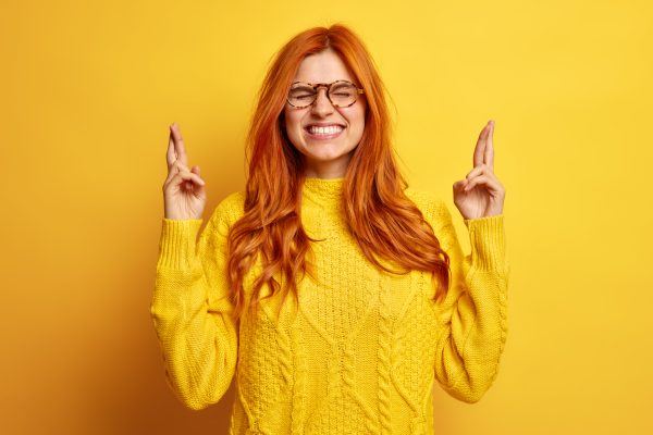Faithful pretty young redhead woman closes eyes keeps fingers crossed makes wish dressed in casual jumper believes in good luck isolated over yellow background waits impatinately for results