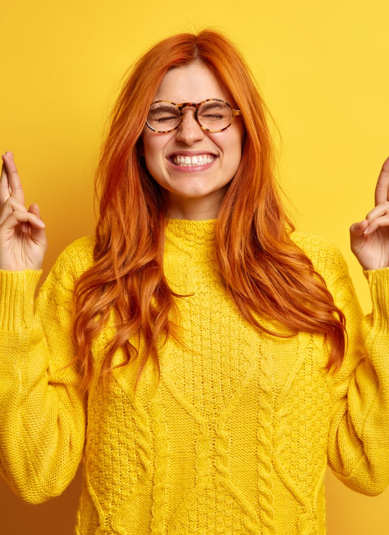 Faithful pretty young redhead woman closes eyes keeps fingers crossed makes wish dressed in casual jumper believes in good luck isolated over yellow background waits impatinately for results