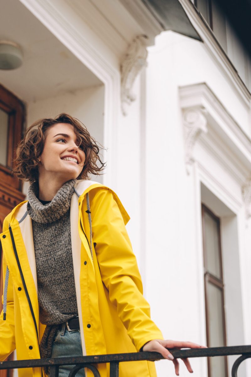 Image of happy pretty young woman dressed in yellow raincoat walking outdoors. Looking aside.