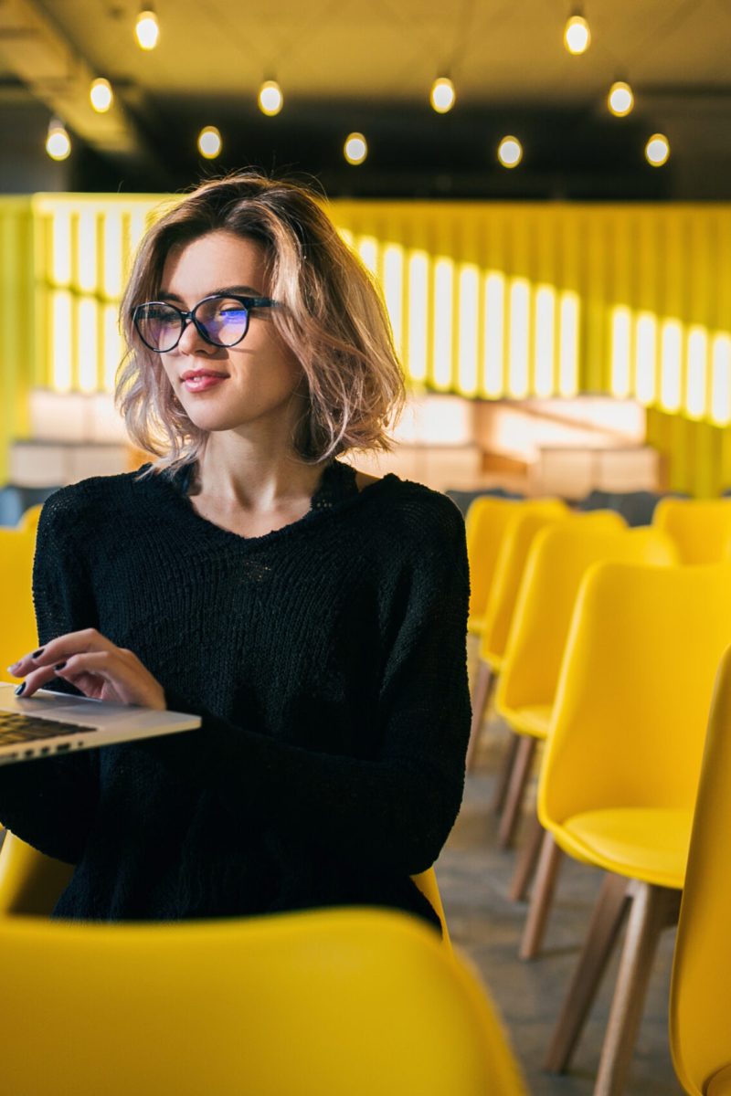 portrait of young attractive woman sitting in lecture hall working on laptop wearing glasses, student learning in classroom with many yellow chairs